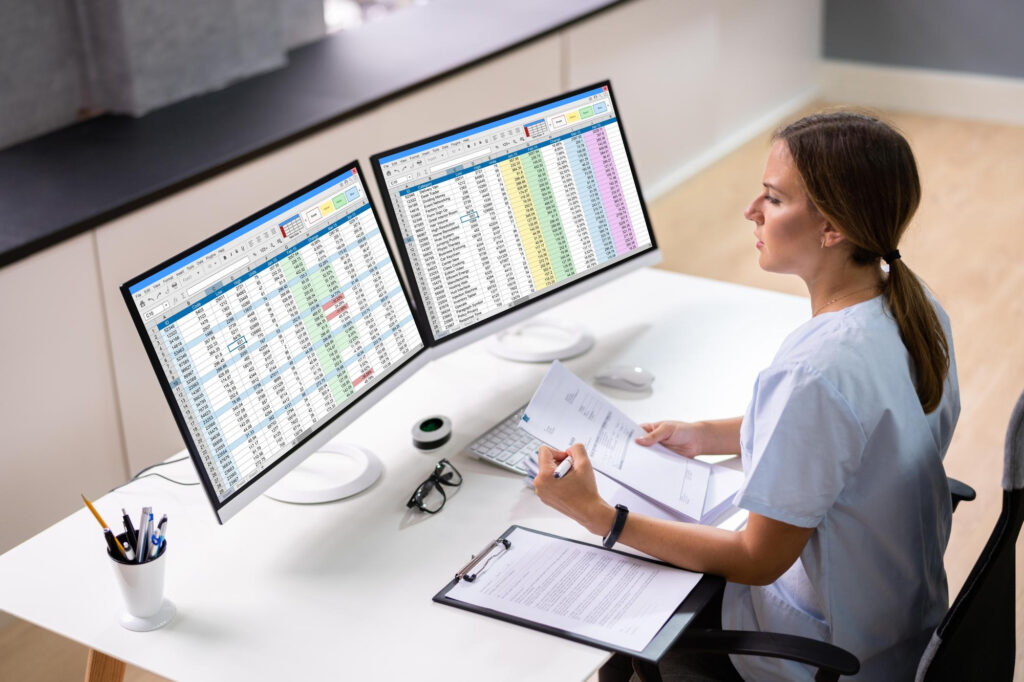 Profesional de la salud gestionando inventario y mantenimiento de equipos médicos mediante hojas de cálculo en doble monitor.